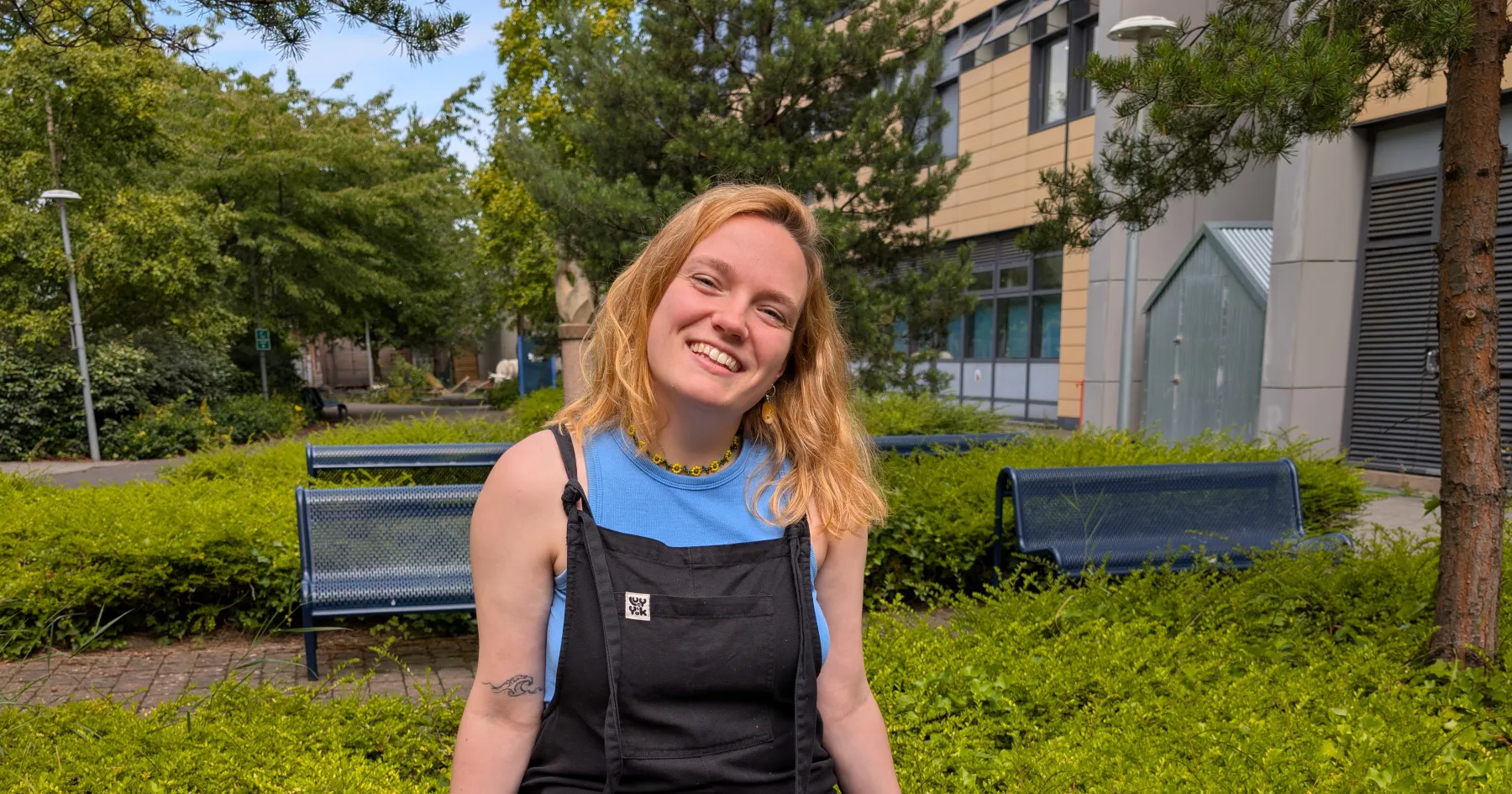 Woman sits on the back of a bench wearing dungarees.